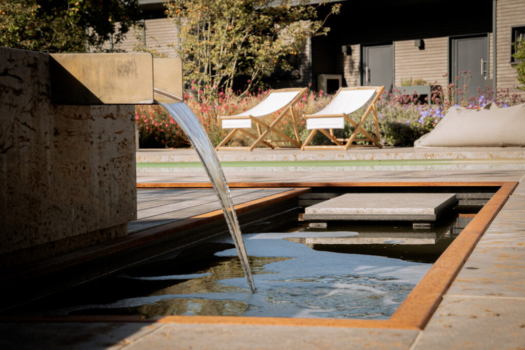 Wasserlauf im Garten. Im Hinetrgrund stehen hochwertige Outdoormöbel am Pool.